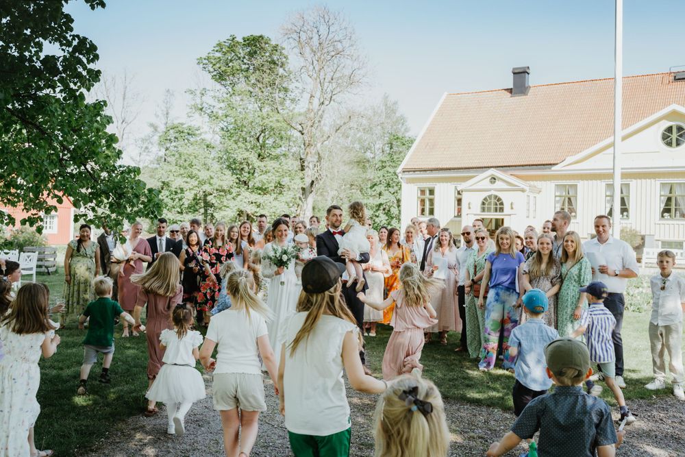 Large group of wedding guests gathered outside on sunny day with white buildings in background.
