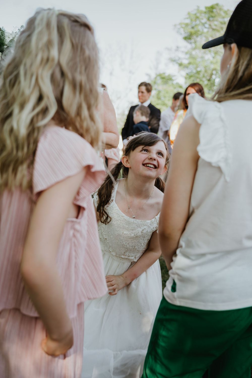 Young attendee in white dress smiles during outdoor wedding celebration.