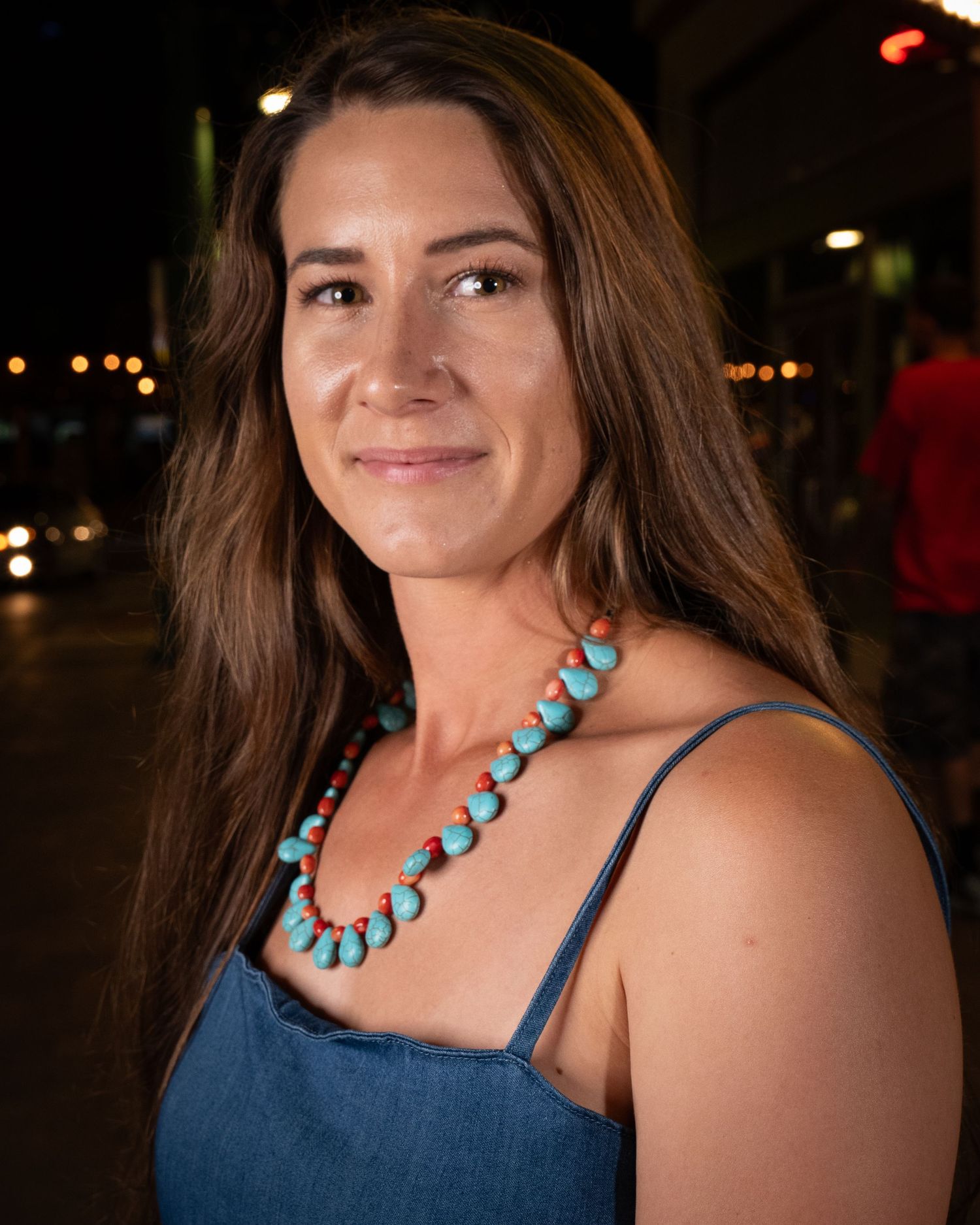 Woman wearing turquoise necklace and denim dress smiling in evening street setting.