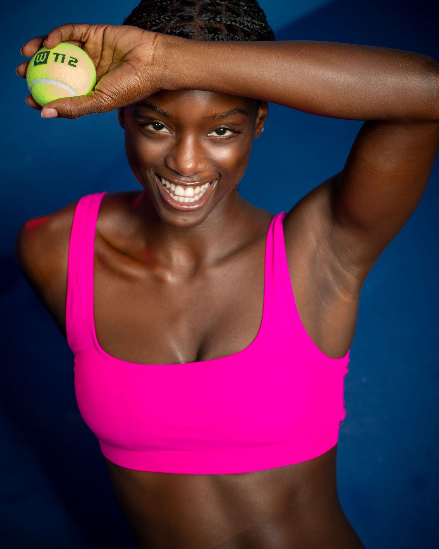 Athletic figure in pink sports bra holds tennis ball against blue background while smiling joyfully.