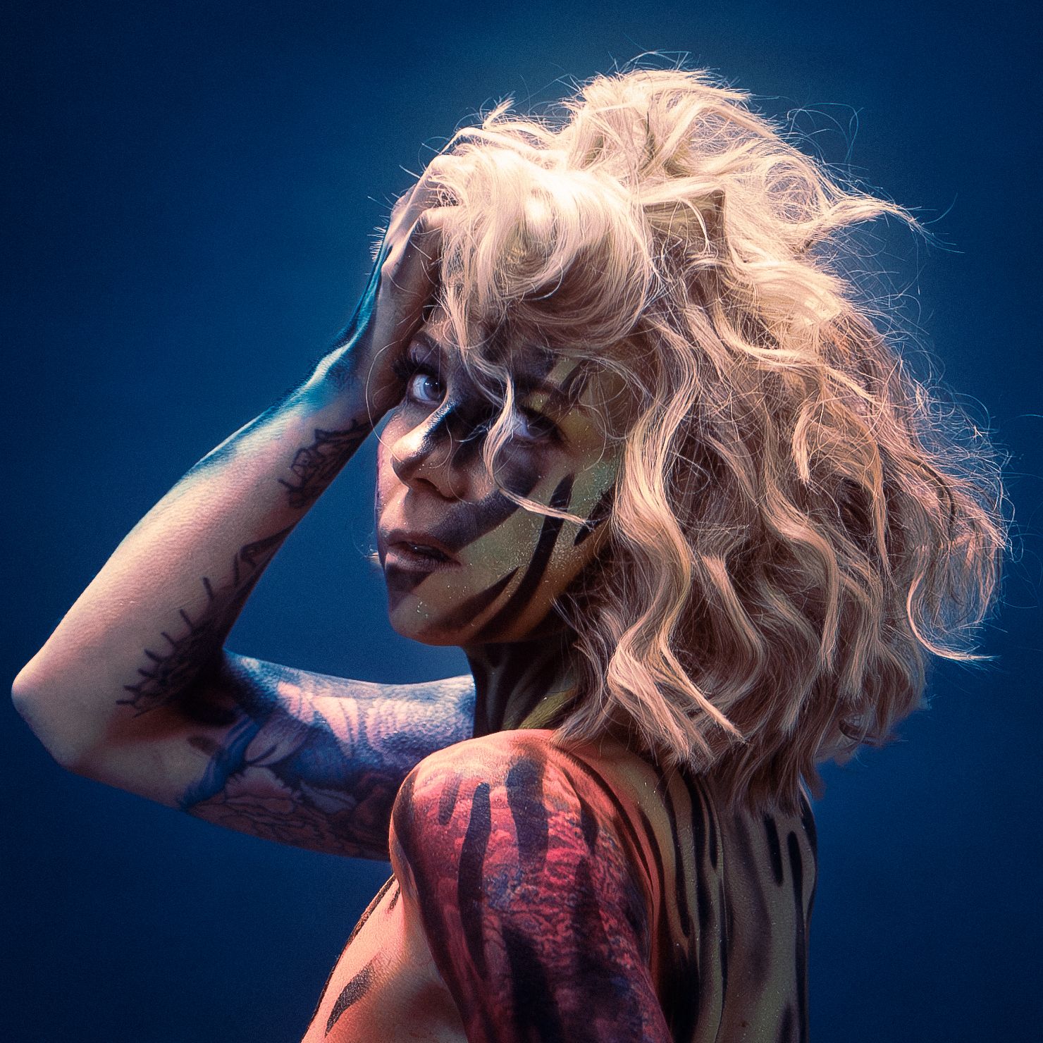 Side profile of a model with flowing blonde hair and artistic body paint against blue background.