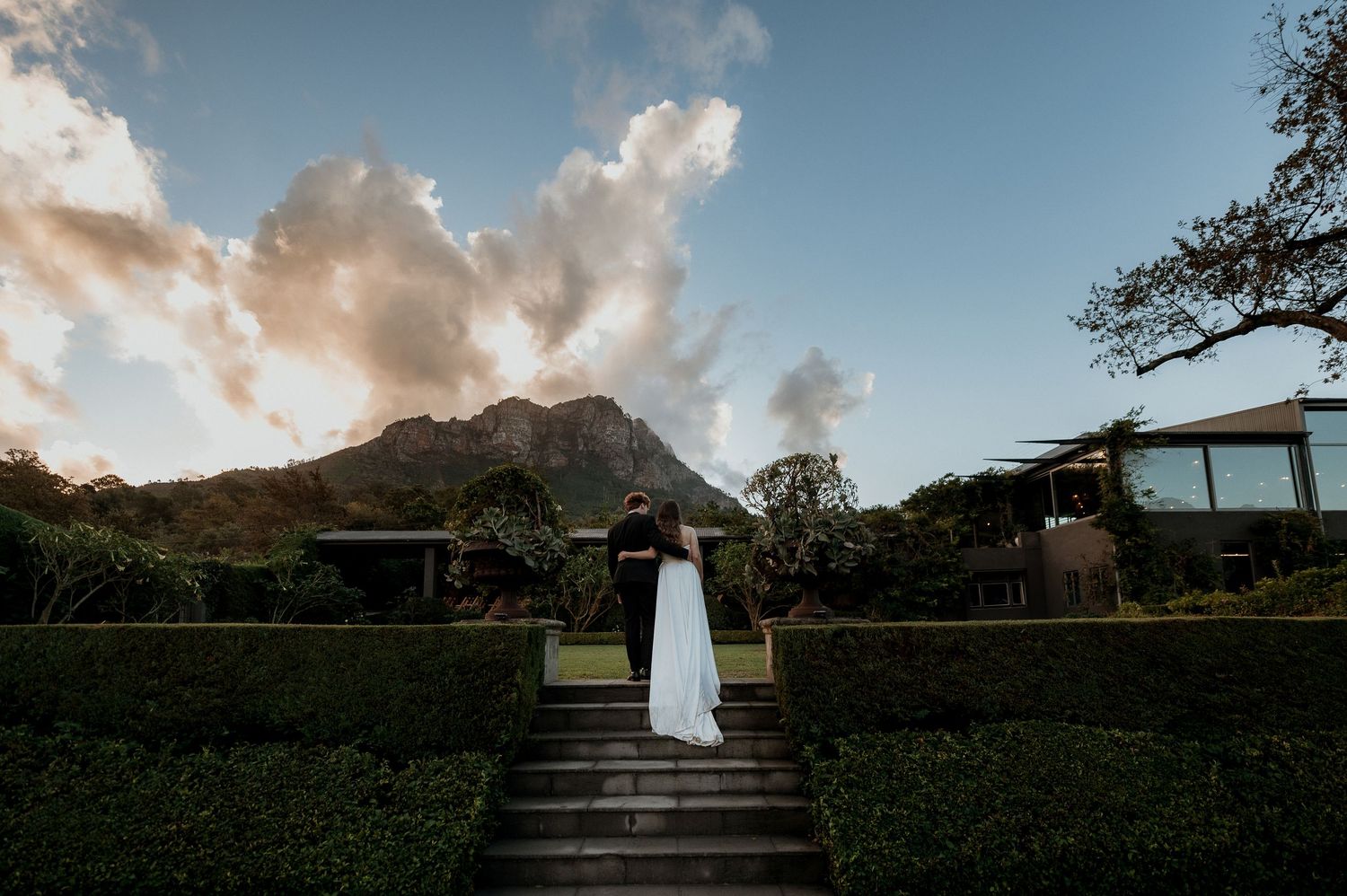 Couple embracing on garden steps with mountain backdrop at sunset, surrounded by lush hedges.