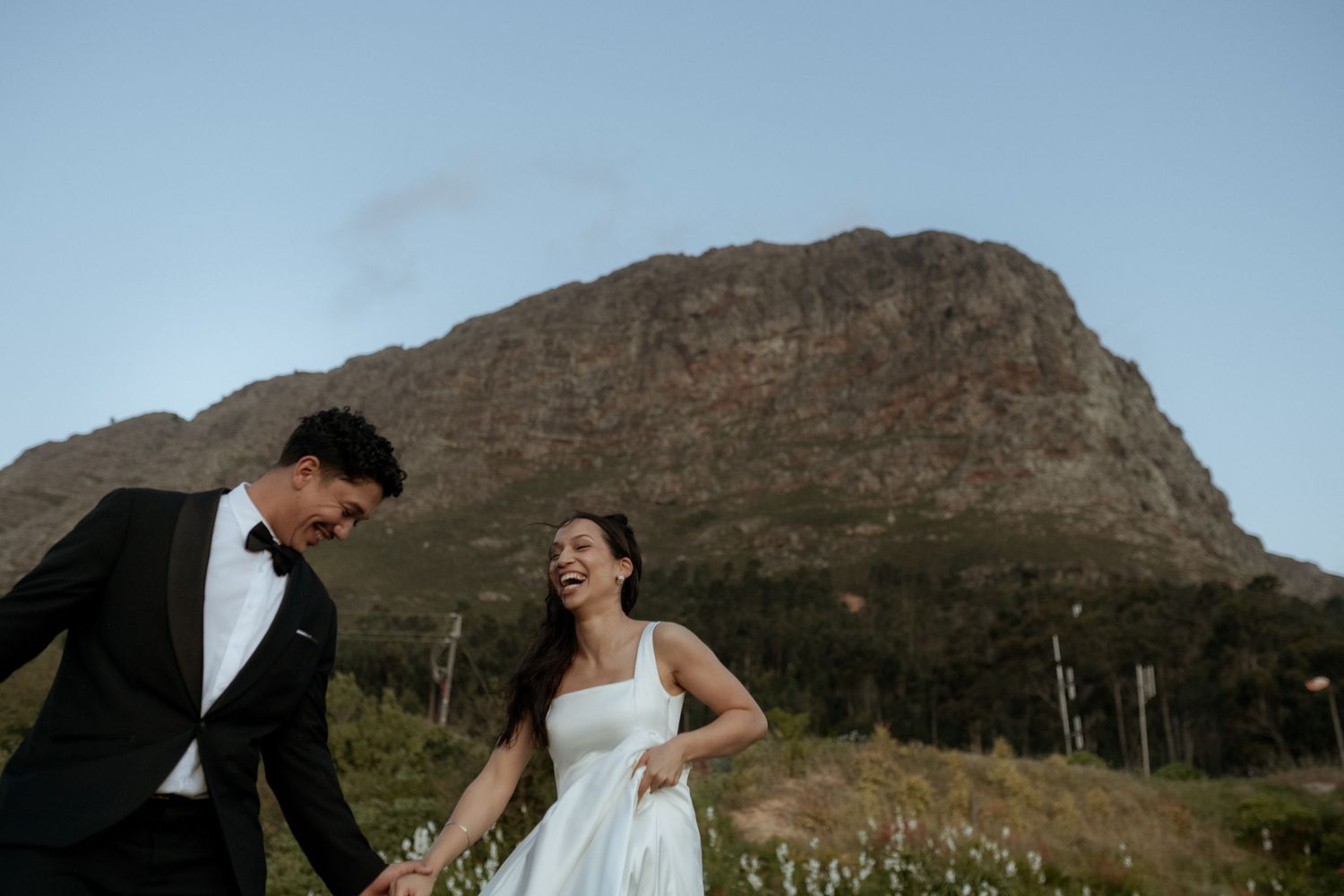 Couple in wedding attire laughing and holding hands in a mountain meadow, bride in white dress, groom in black tuxedo.