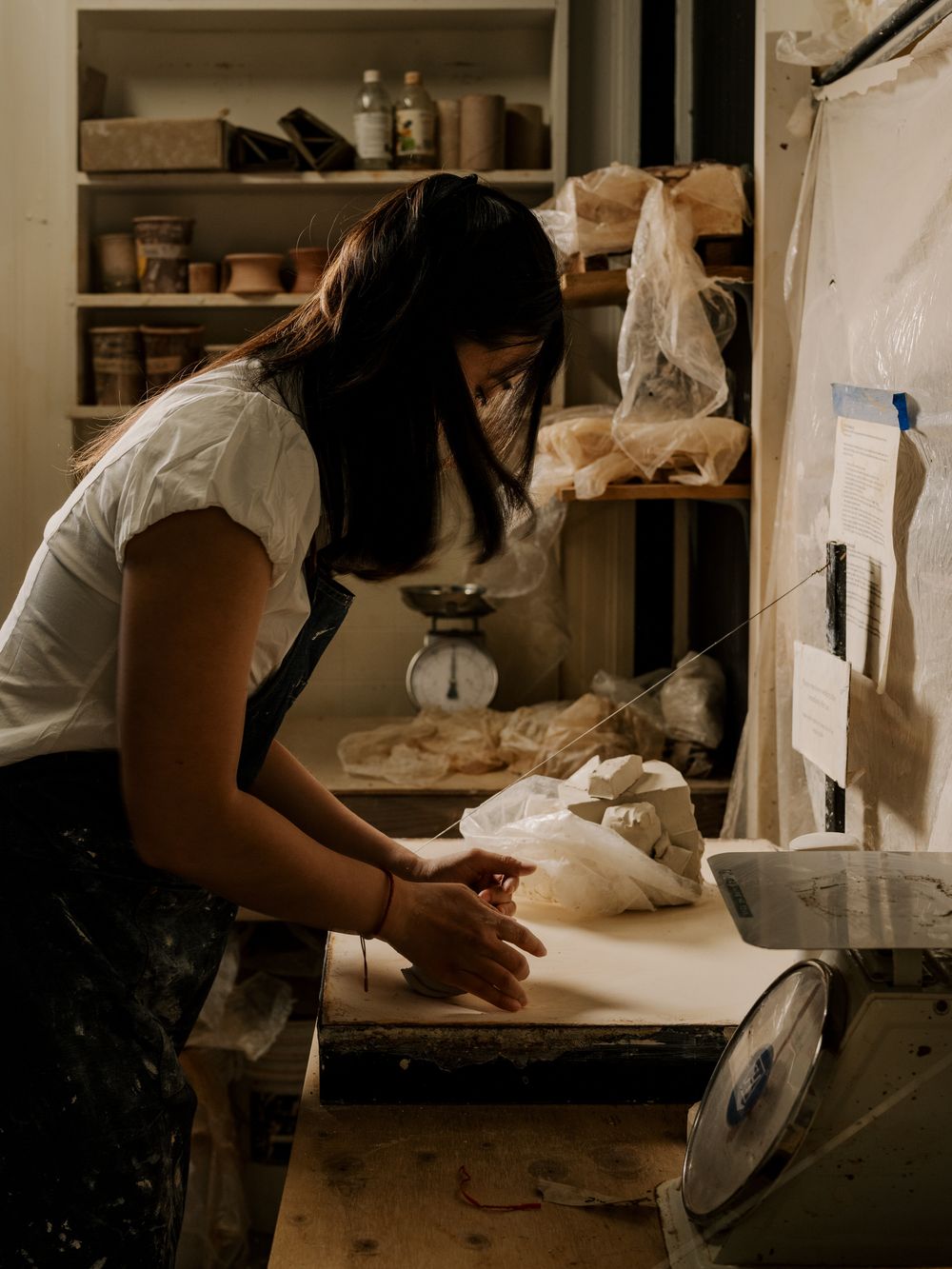 Ceramics studio workspace shows hands working with clay on a table surrounded by shelves of pottery tools and supplies.