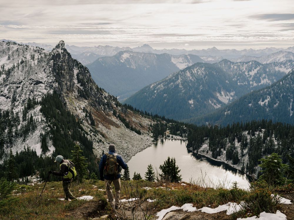 Hikers stand on a mountain ridge overlooking a dramatic landscape of snow-dusted peaks and valleys in the wilderness.