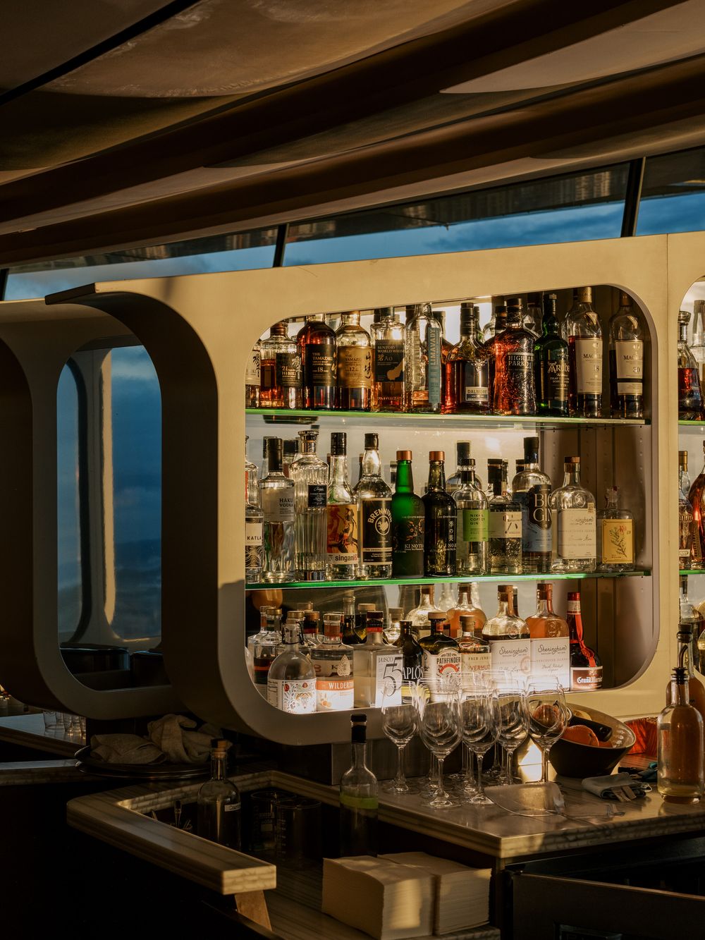 A well-stocked bar shelf with various spirits and wine glasses displayed through a modern porthole-style window frame.