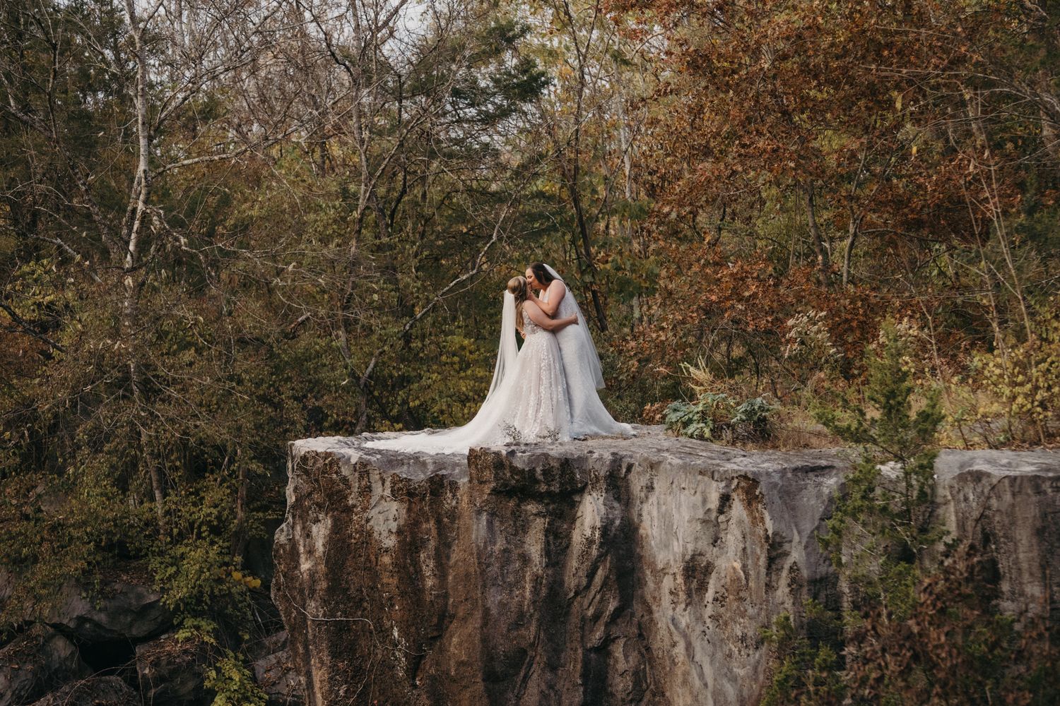 A bride in white dress sits on stone wall surrounded by autumn trees.