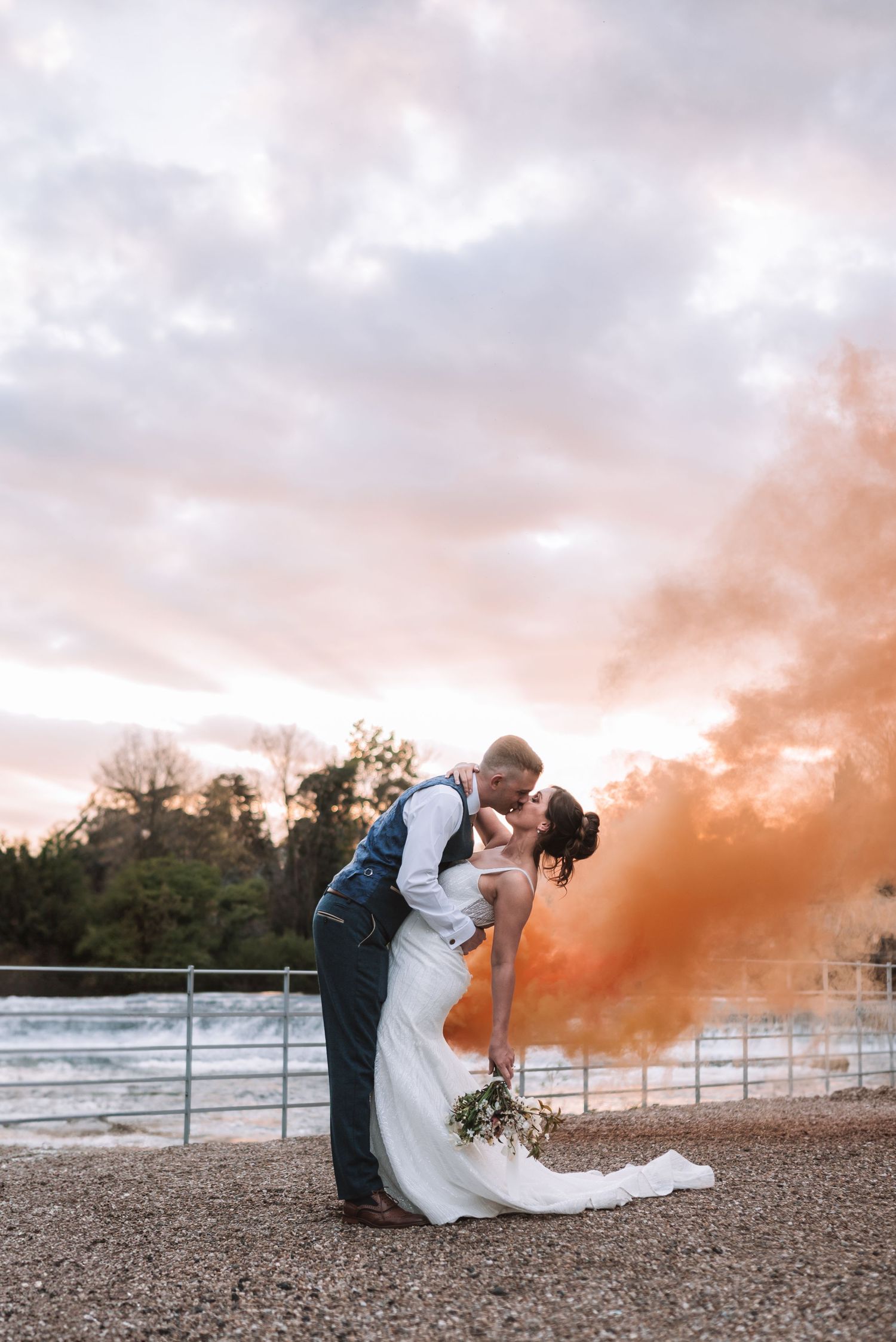 A romantic couple embraces against a sunset sky with orange smoke bombs creating a dramatic atmospheric effect at the West Mill.