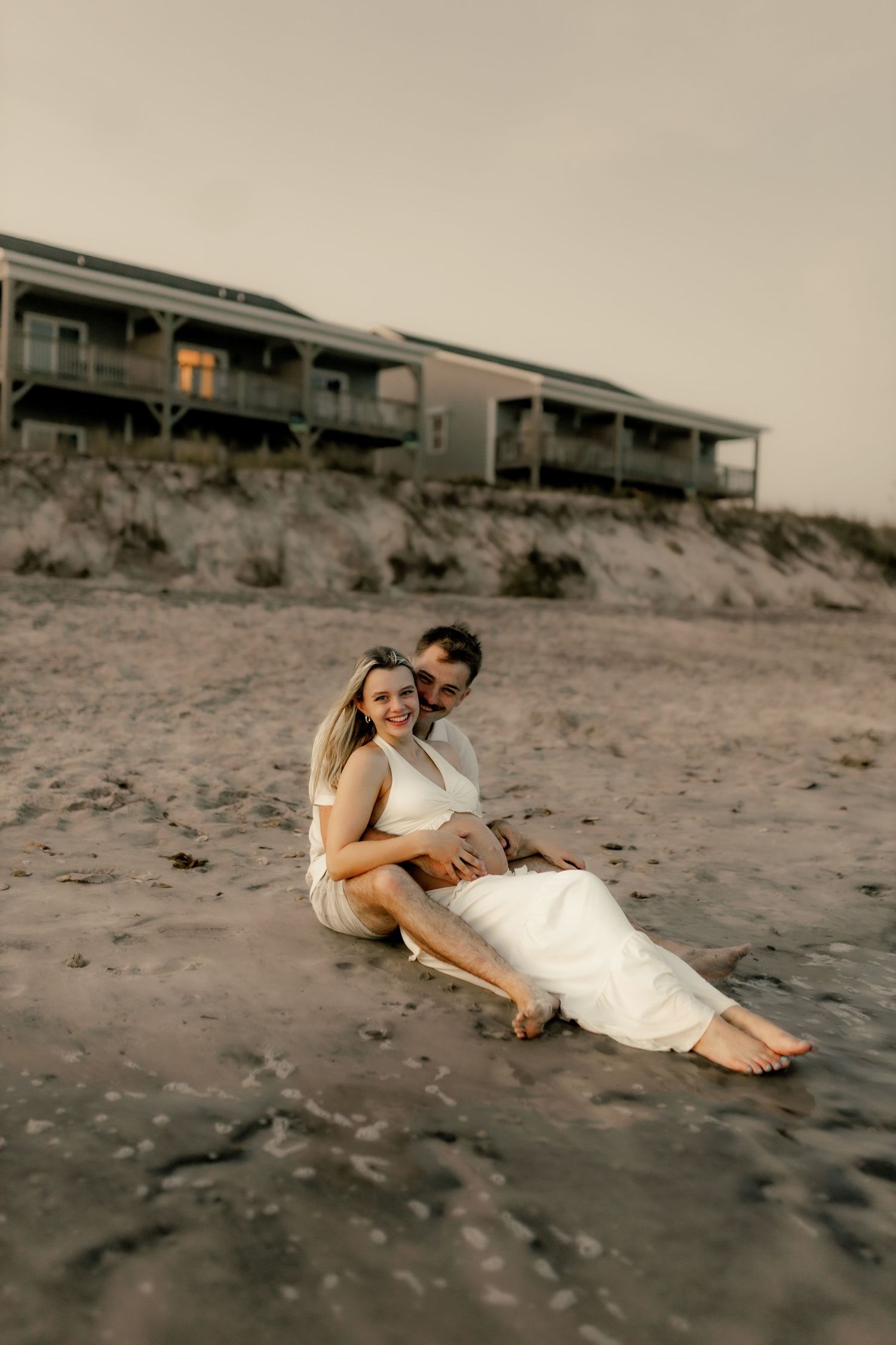 Couple in white outfits sharing intimate moments on beach with beach houses in background.