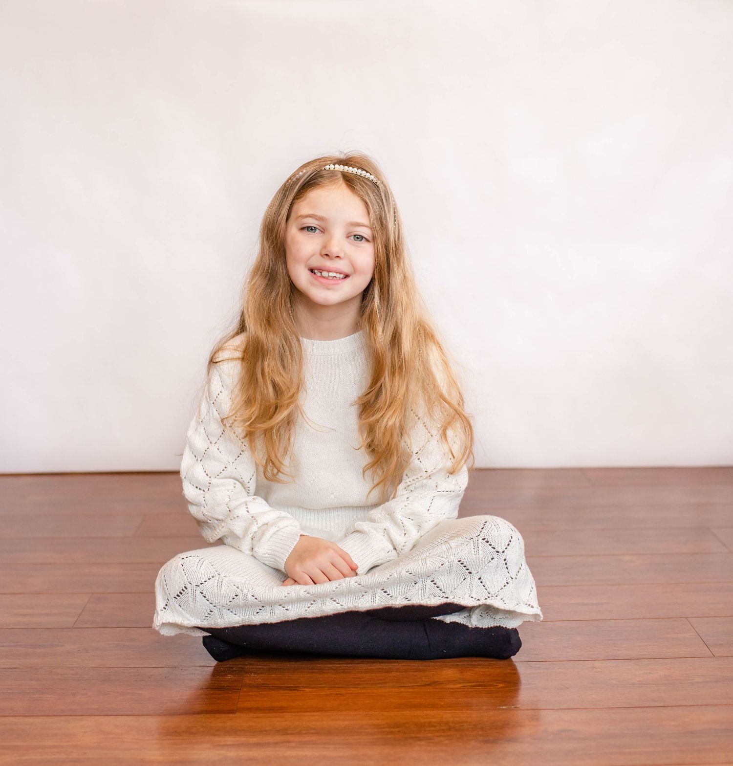 Young person in white sweater sits cross-legged on wooden floor with bright smile.