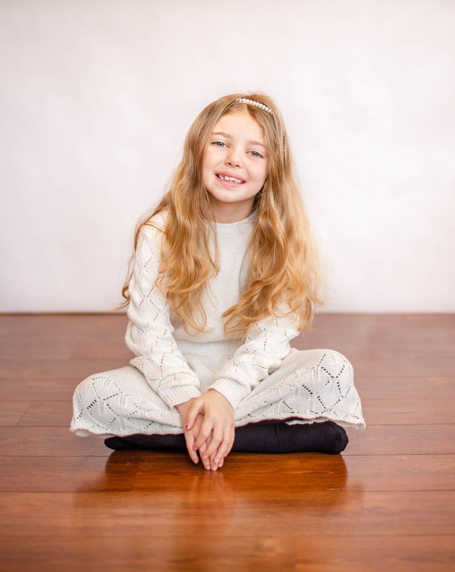 Young person in cozy white sweater sits cross-legged on hardwood floor.