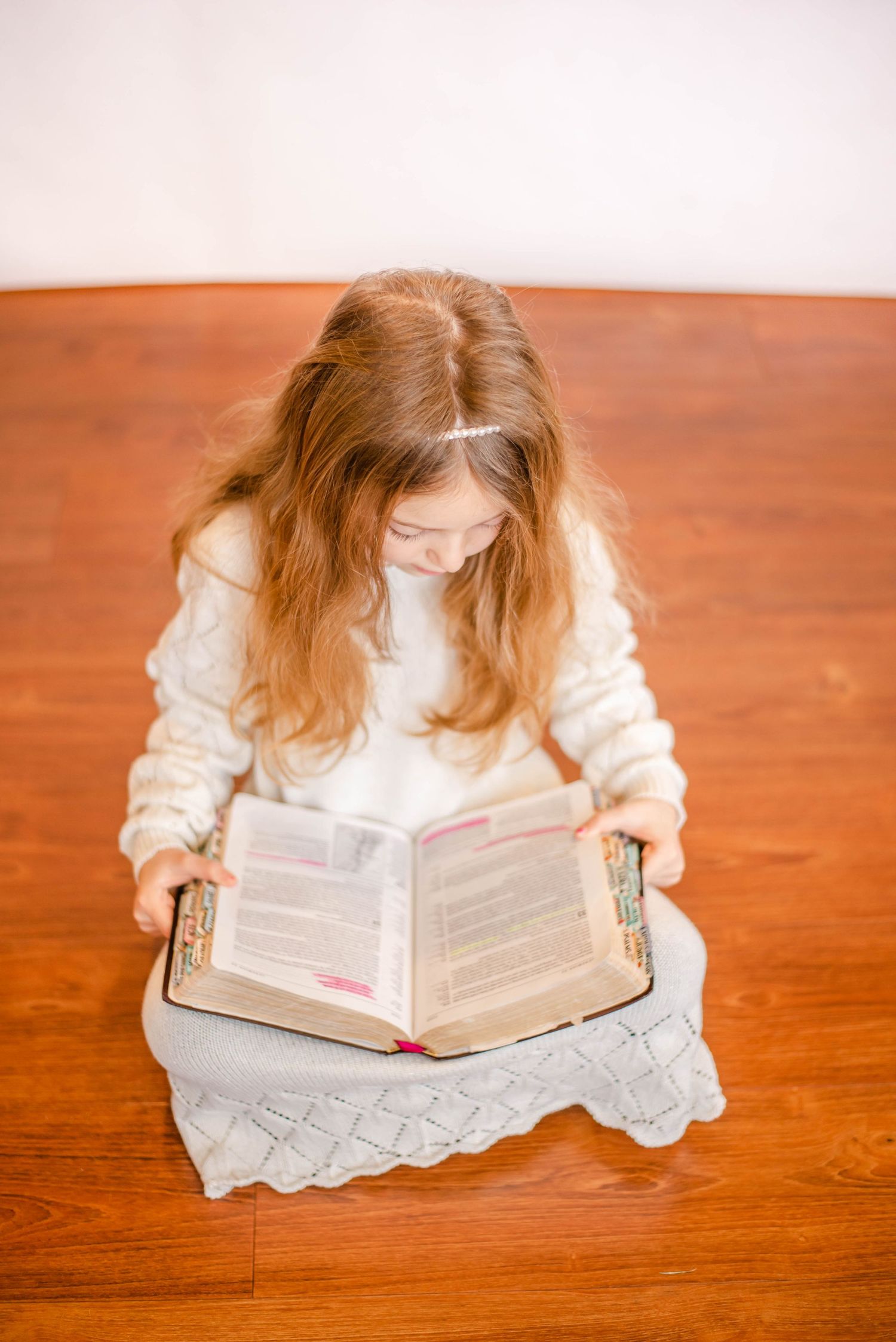 Person with long hair reads intently from book while sitting on wooden floor.