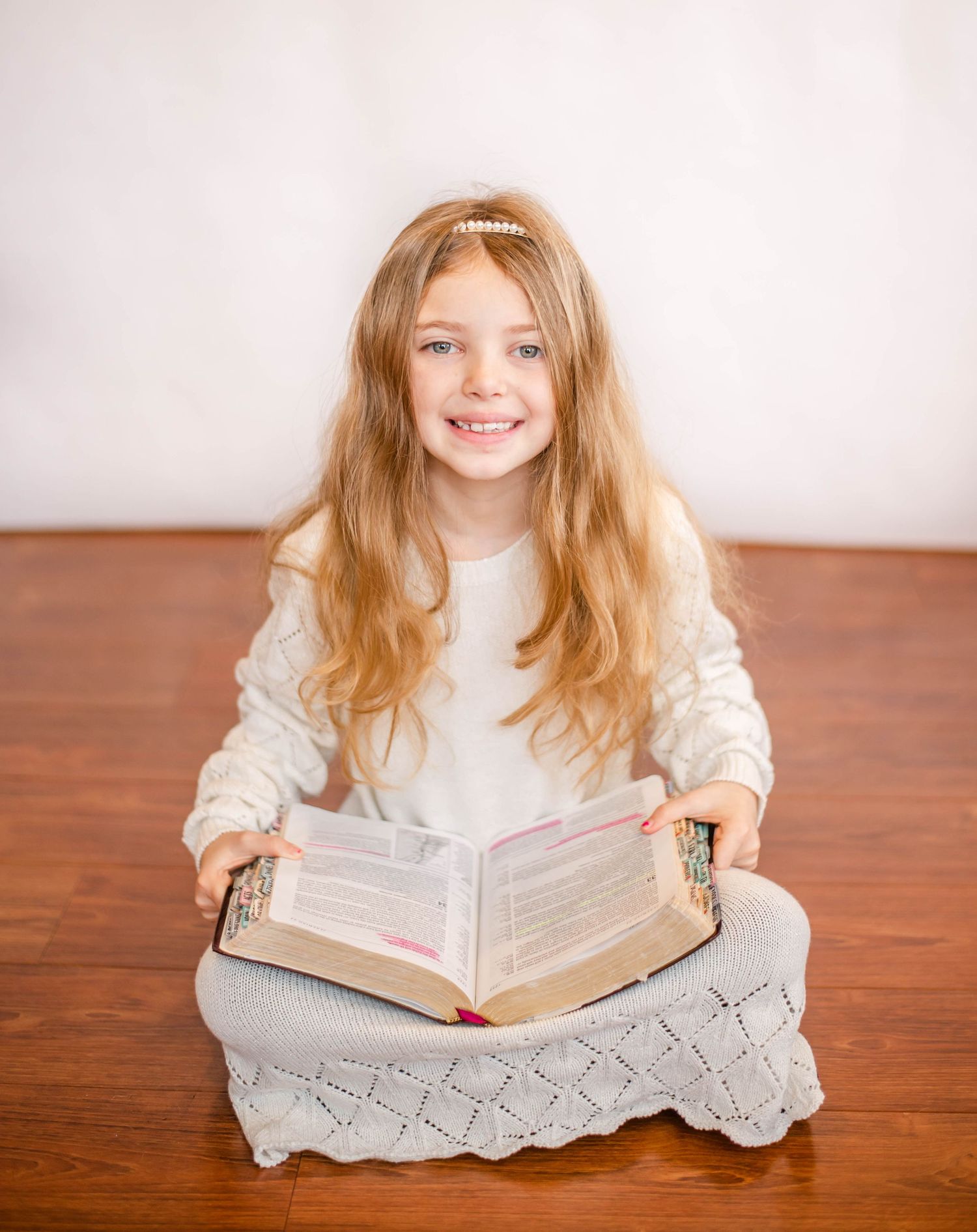 Person in white sweater smiles while holding open book on wooden floor.