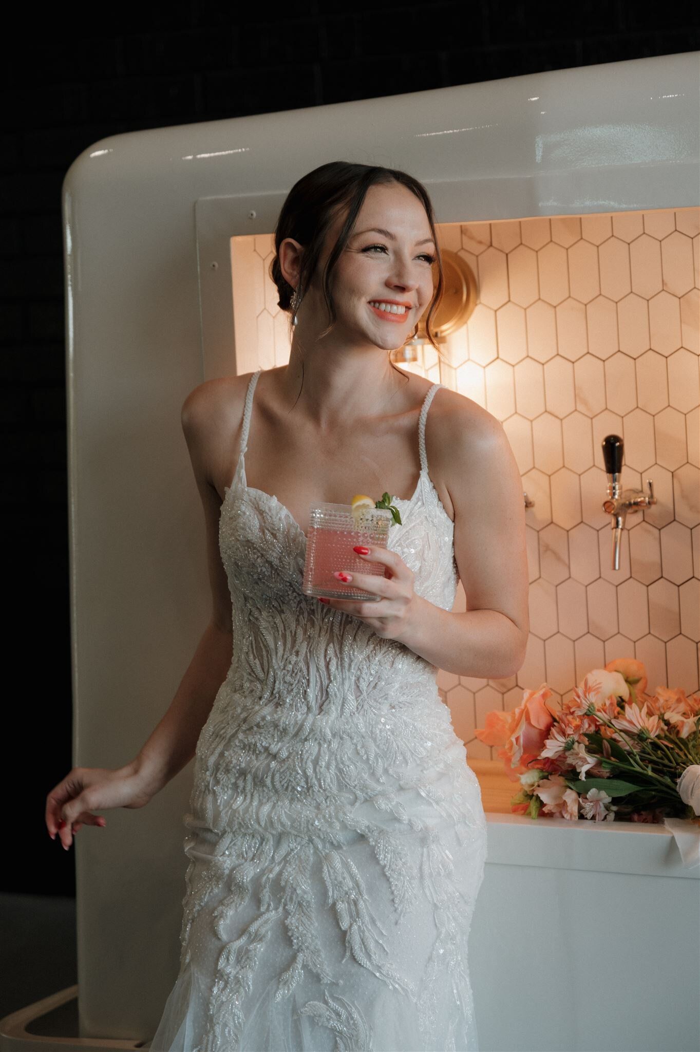 A bride in a stunning lace wedding dress smiles while holding a pink drink in a modern room with honeycomb tile.