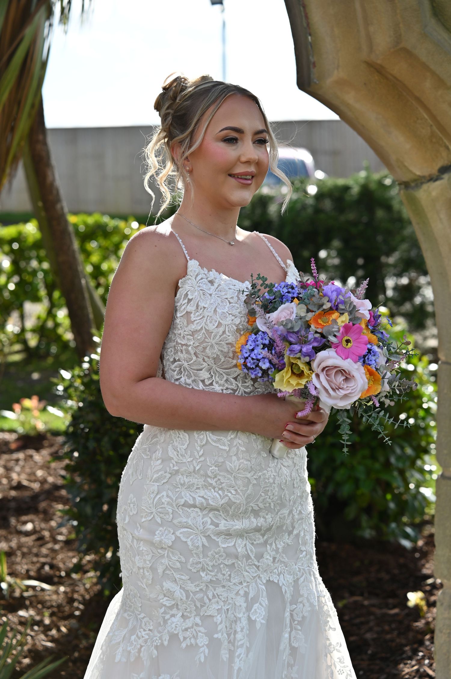 A bride in a white lace wedding dress holds a vibrant bouquet of purple, pink and yellow flowers in a garden setting.