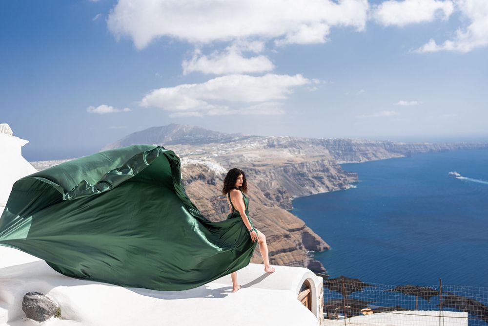 A flowing green dress billows dramatically against the backdrop of Santorini's caldera and white buildings.
