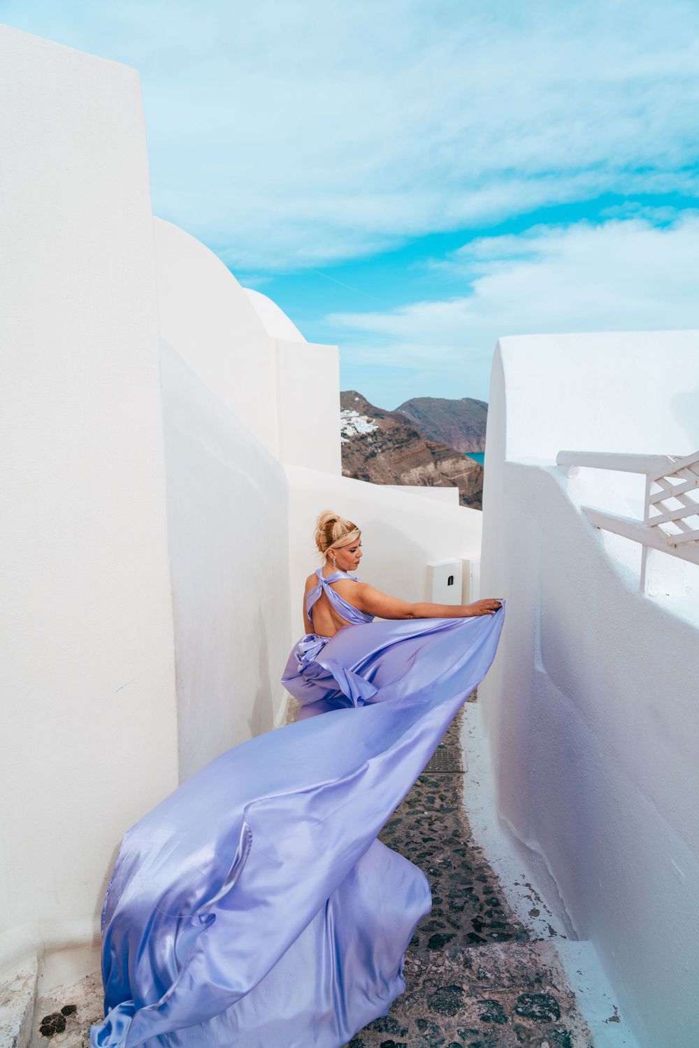 A lavender dress trails elegantly through a white-walled Santorini pathway with mountain views.