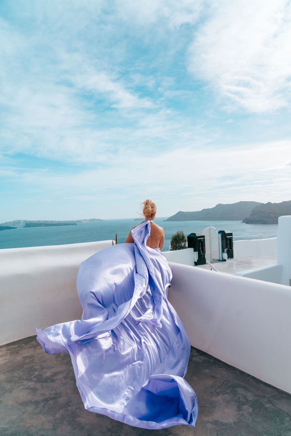 A flowing lavender dress billows dramatically against the backdrop of Santorini's white buildings and blue sea.
