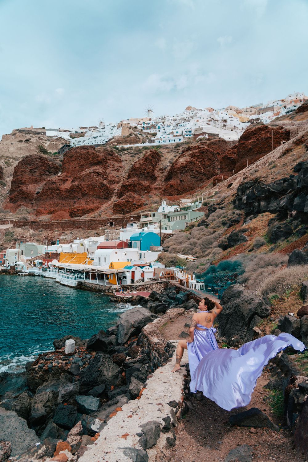 A lavender dress flows in the wind above Santorini's colorful port and red cliffs.