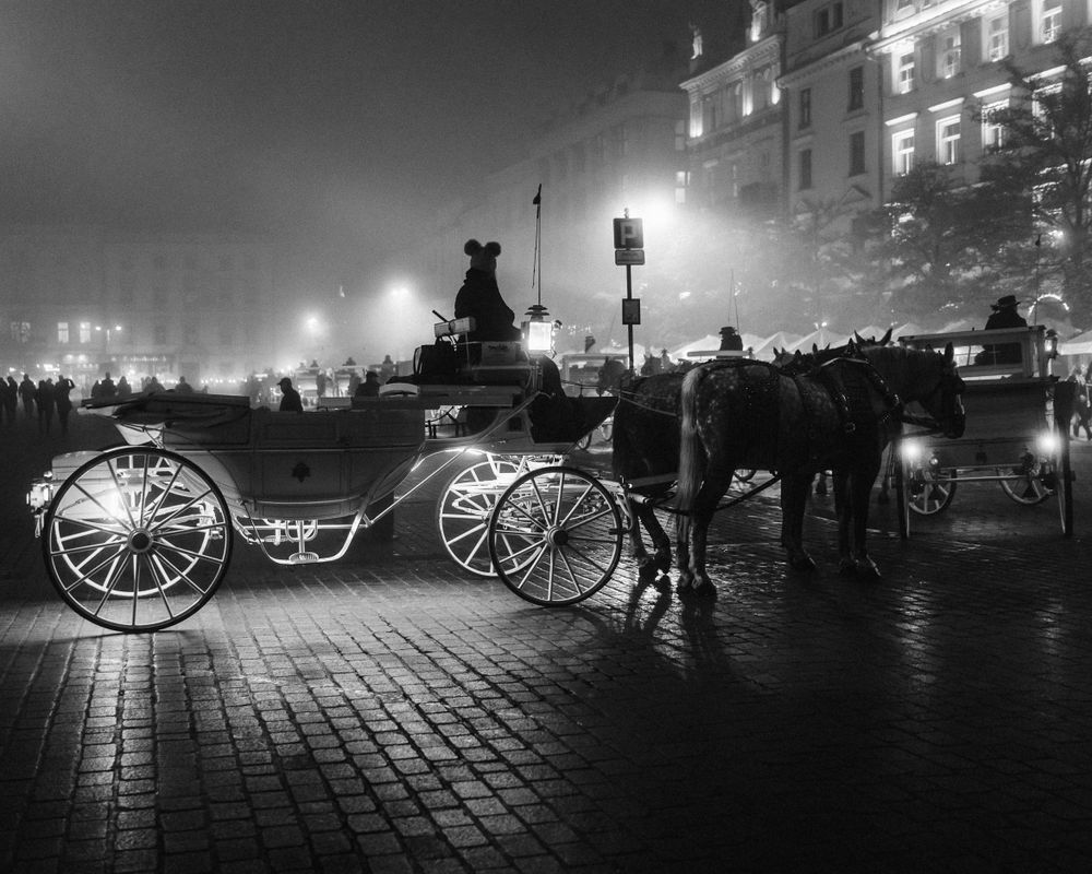 Foggy night scene with horse carriages silhouetted against city lights.