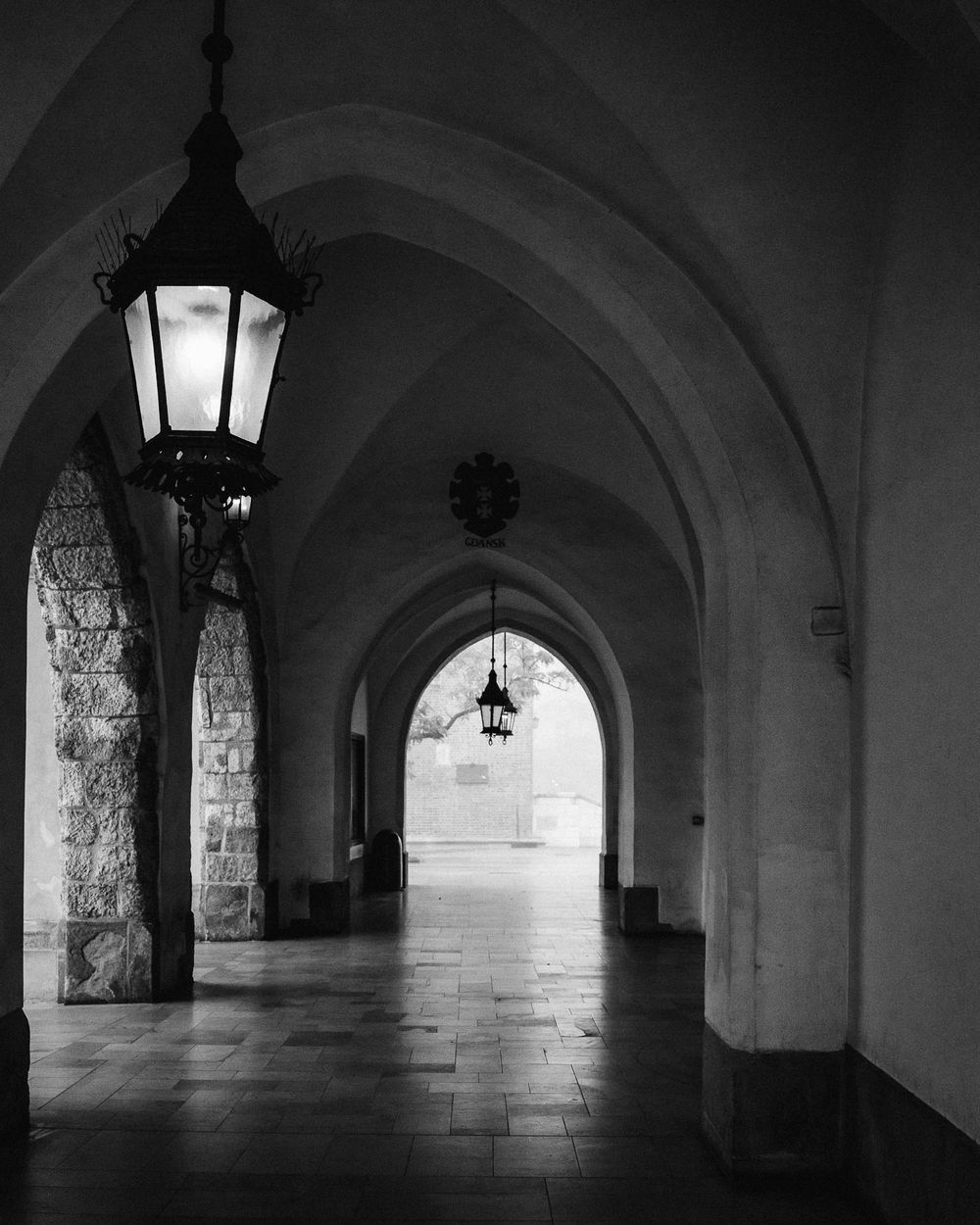 Black and white photo of a medieval-style arched hallway, featuring stone columns and hanging lanterns. The corridor ext