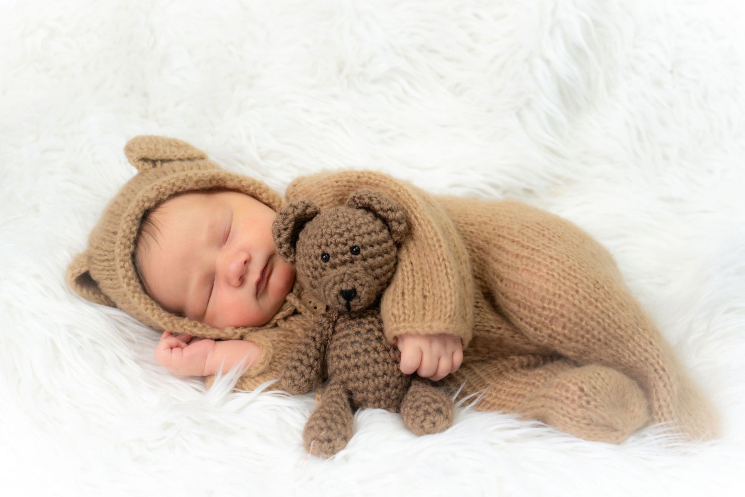 Sleeping newborn in brown knit outfit cuddling with plush teddy bear on white fur background.