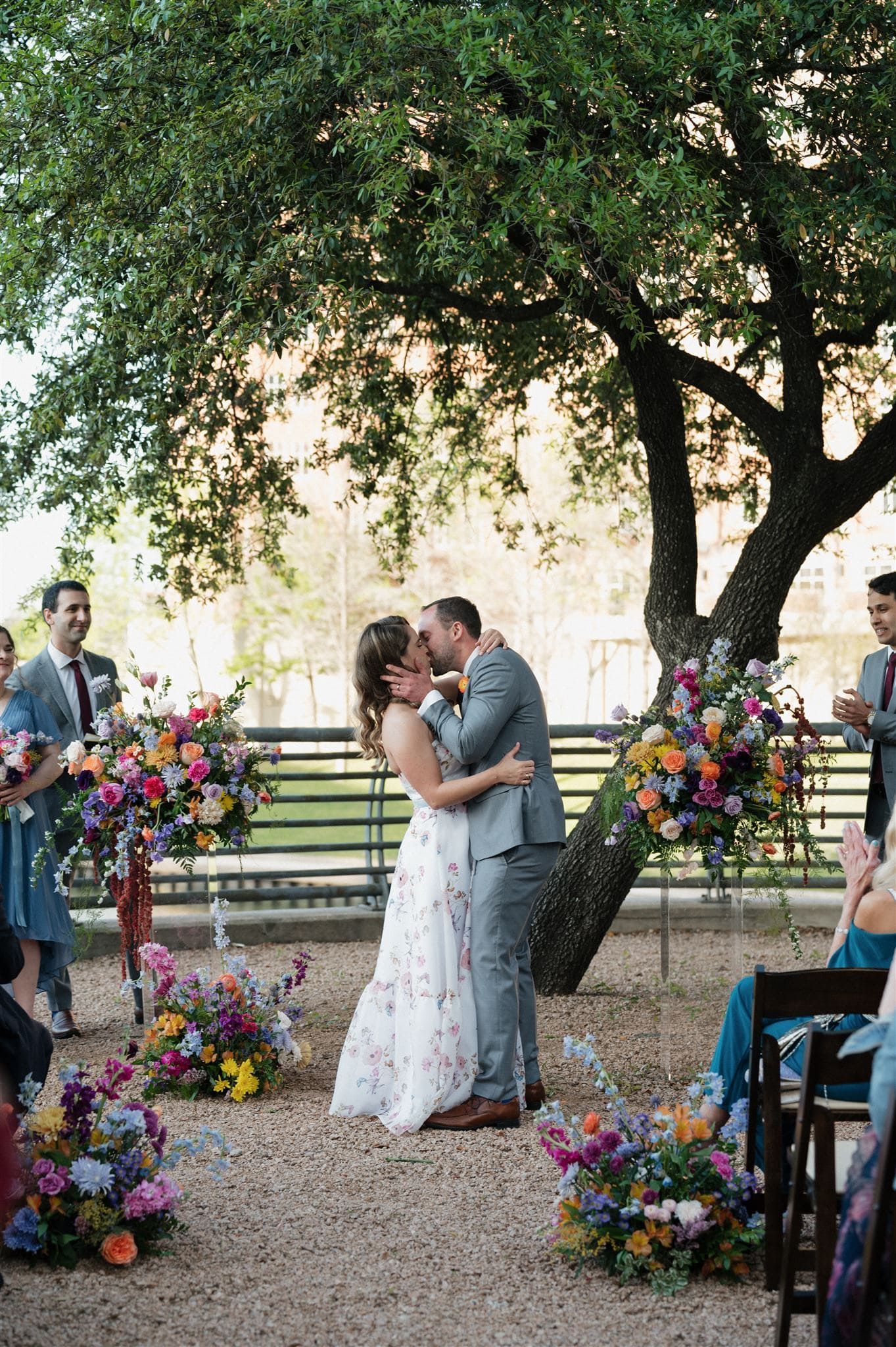 Couple sharing a kiss under a tree surrounded by vibrant floral arrangements at an outdoor wedding.