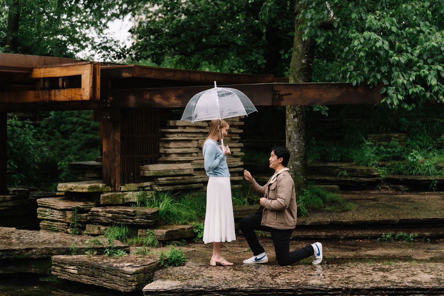 A man kneels on one knee, proposing to a woman holding a clear umbrella on rustic stone steps in a lush, rainy forest setting.