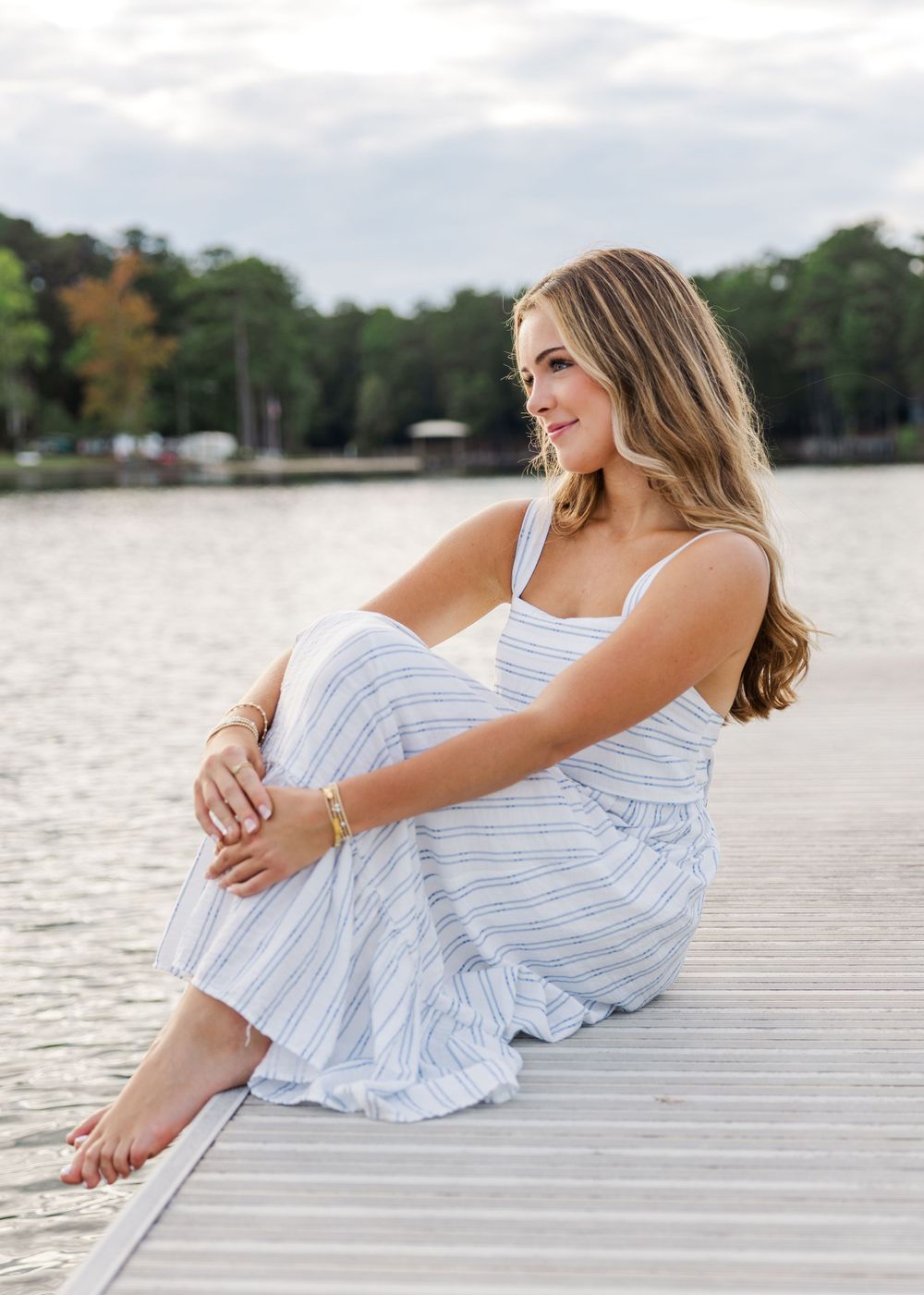 Relaxed pose in blue striped sundress on wooden dock by lake.