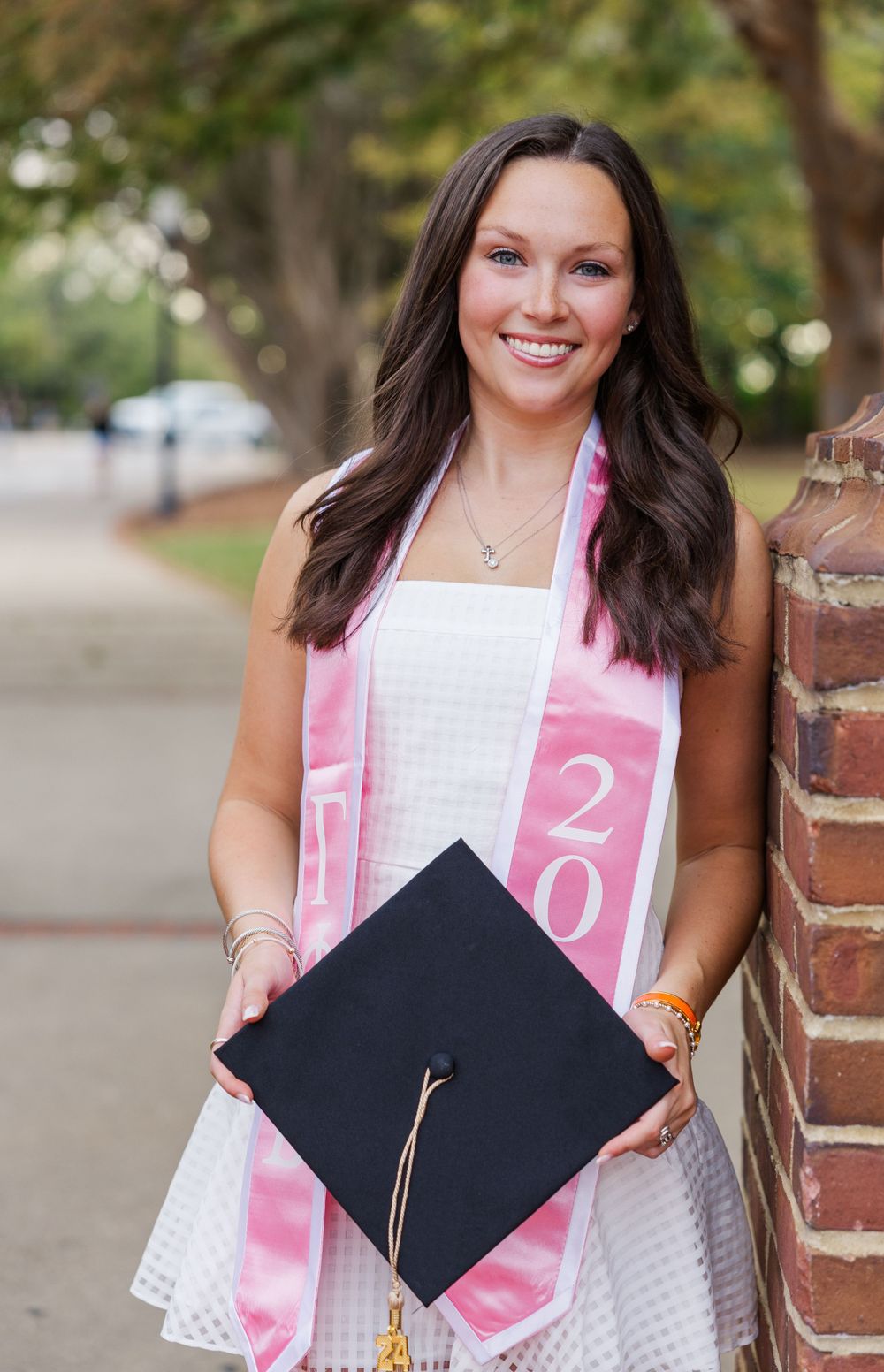 Auburn Graduate in white dress and pink sash holds graduation cap near Samford Lawn by Preslee McGough Photography
