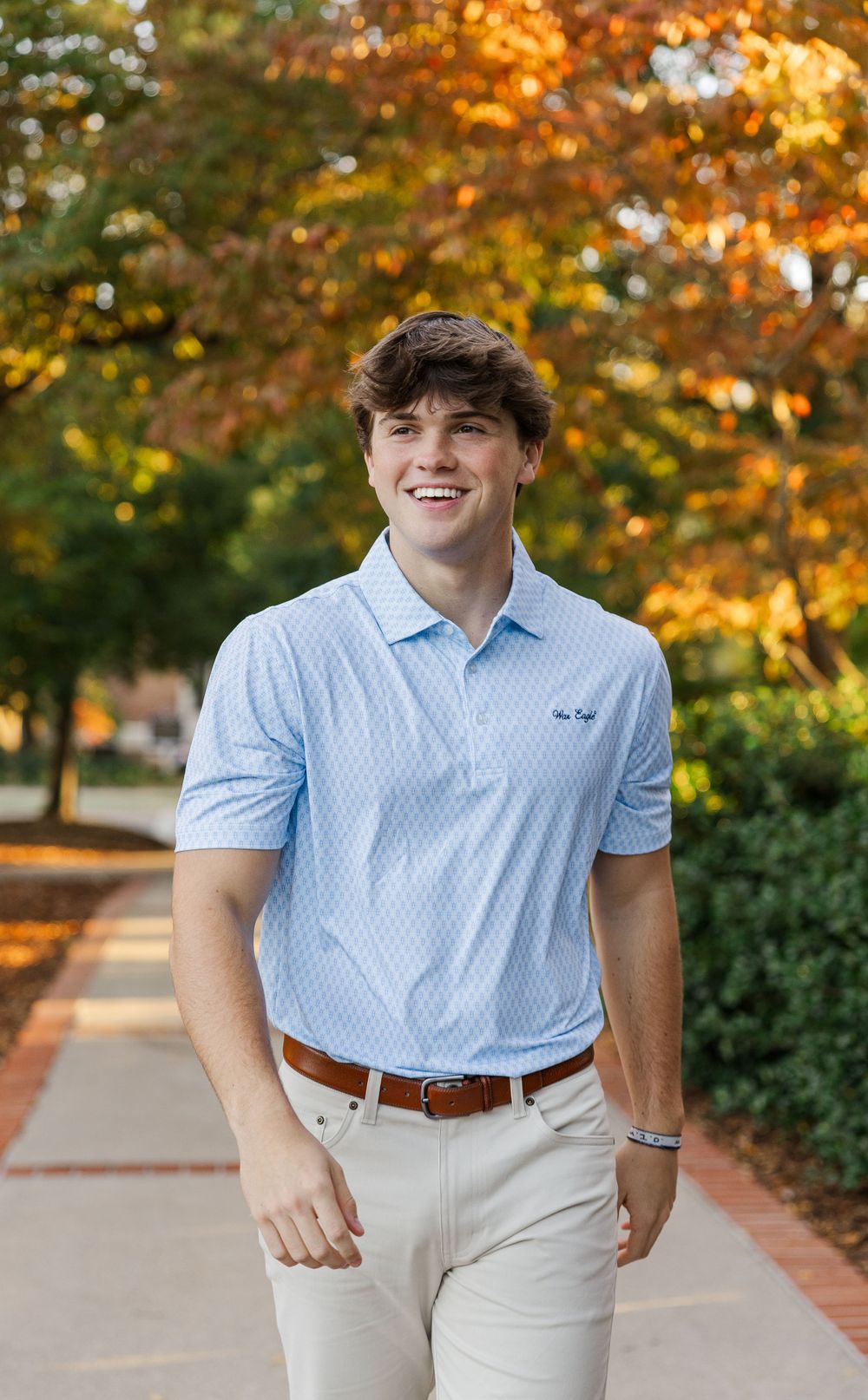 Person in light blue polo shirt and khaki pants smiling outdoors with fall foliage background.