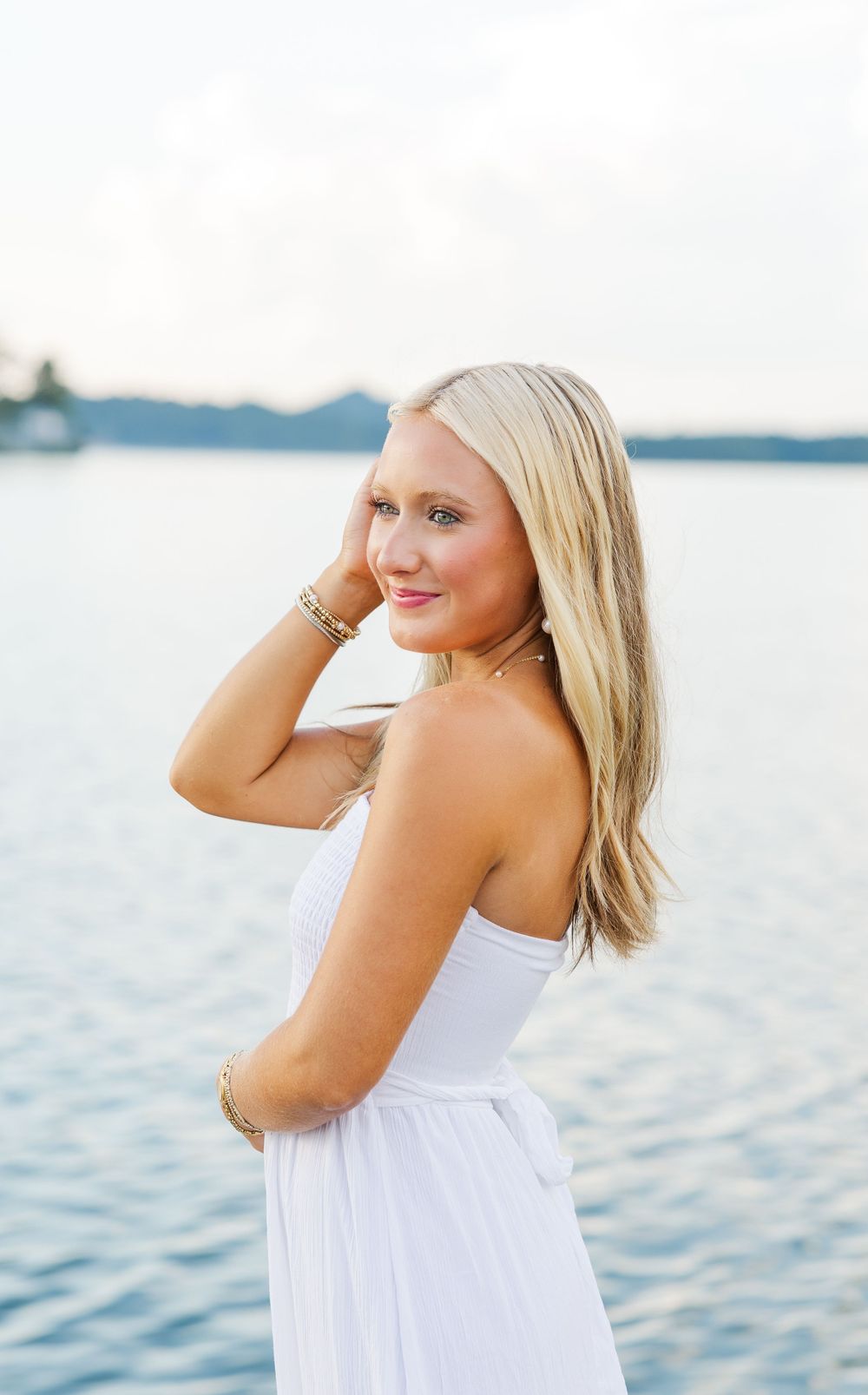 A person in a white sundress poses by a waterfront during sunset.