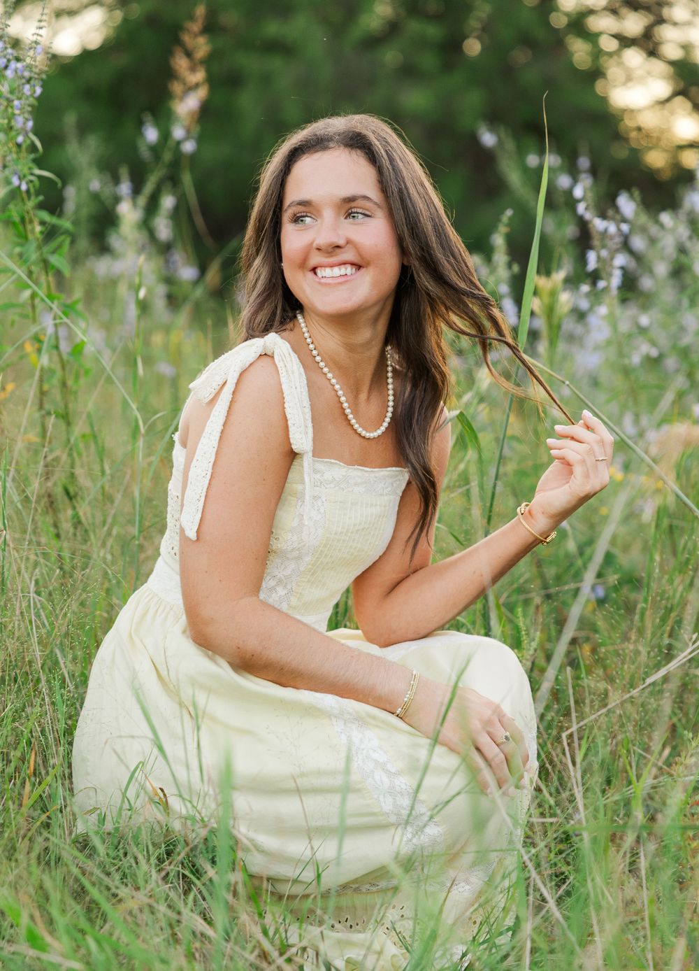 A person in a cream-colored dress poses gracefully in a field of tall grass and wildflowers at sunset.