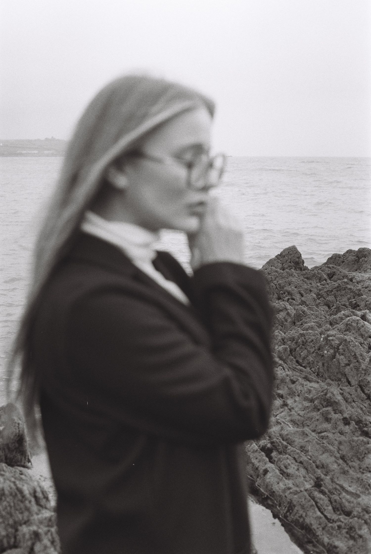 Person in glasses and dark coat poses thoughtfully by rocky ocean shore in soft focus.