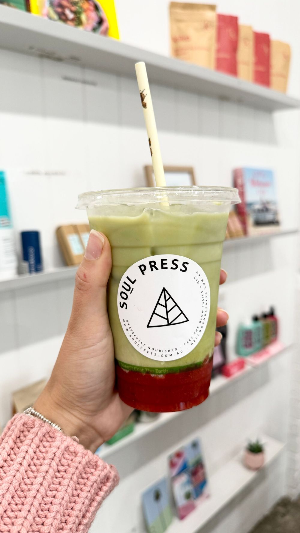 Hand holding a Soul Press branded drink with matcha and red layers against shelves of products.