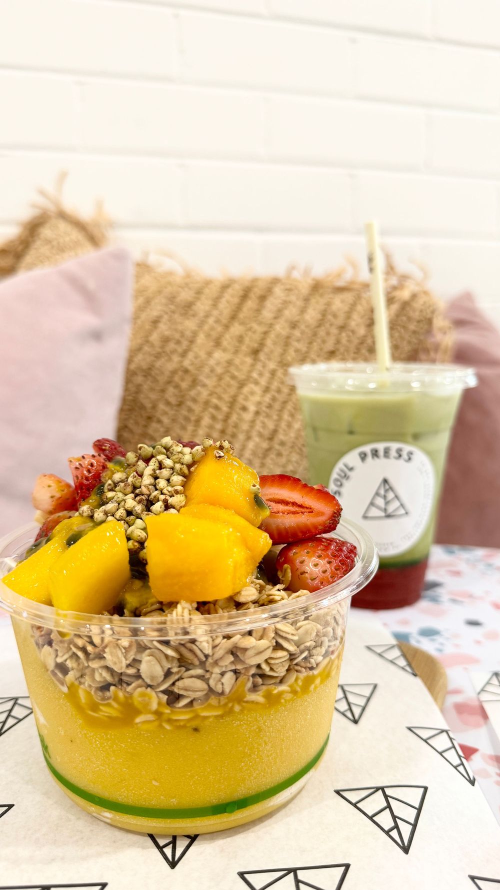 Fresh fruit bowl with mango and granola next to green Soul Press smoothie on patterned surface.