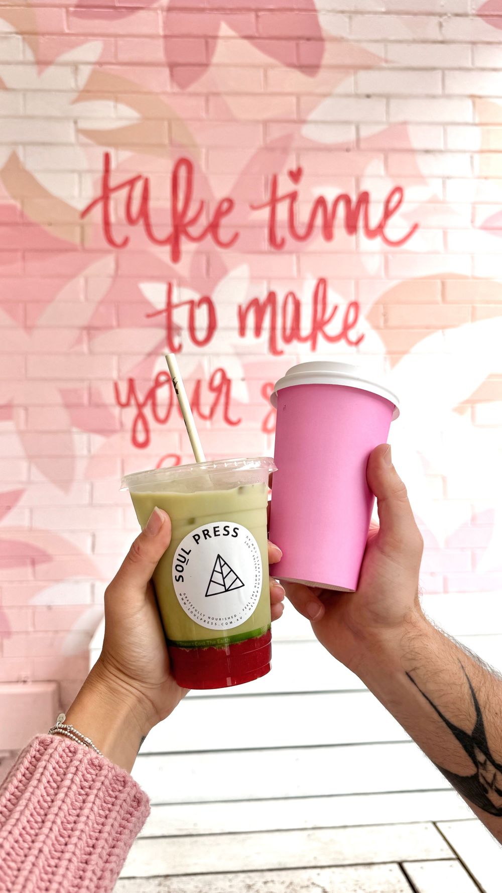 Two hands holding Soul Press drink and pink cup against floral wall art.