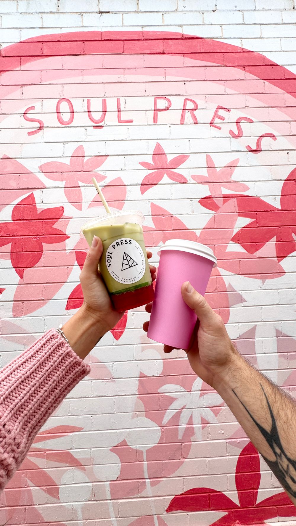 Hands holding Soul Press drink and pink cup against floral mural wall with logo.