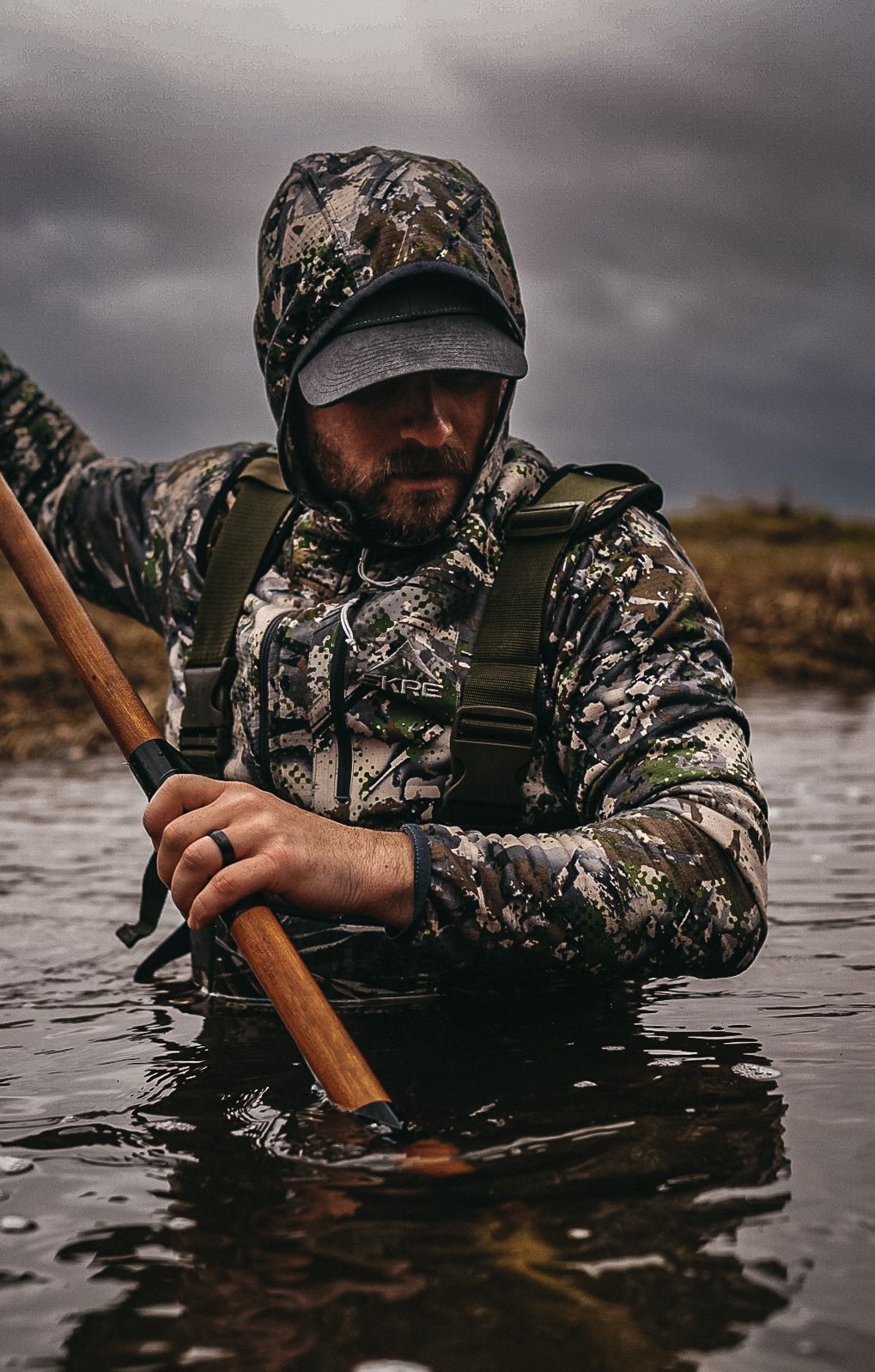 Hunter in camouflage gear wading through water with hunting equipment.