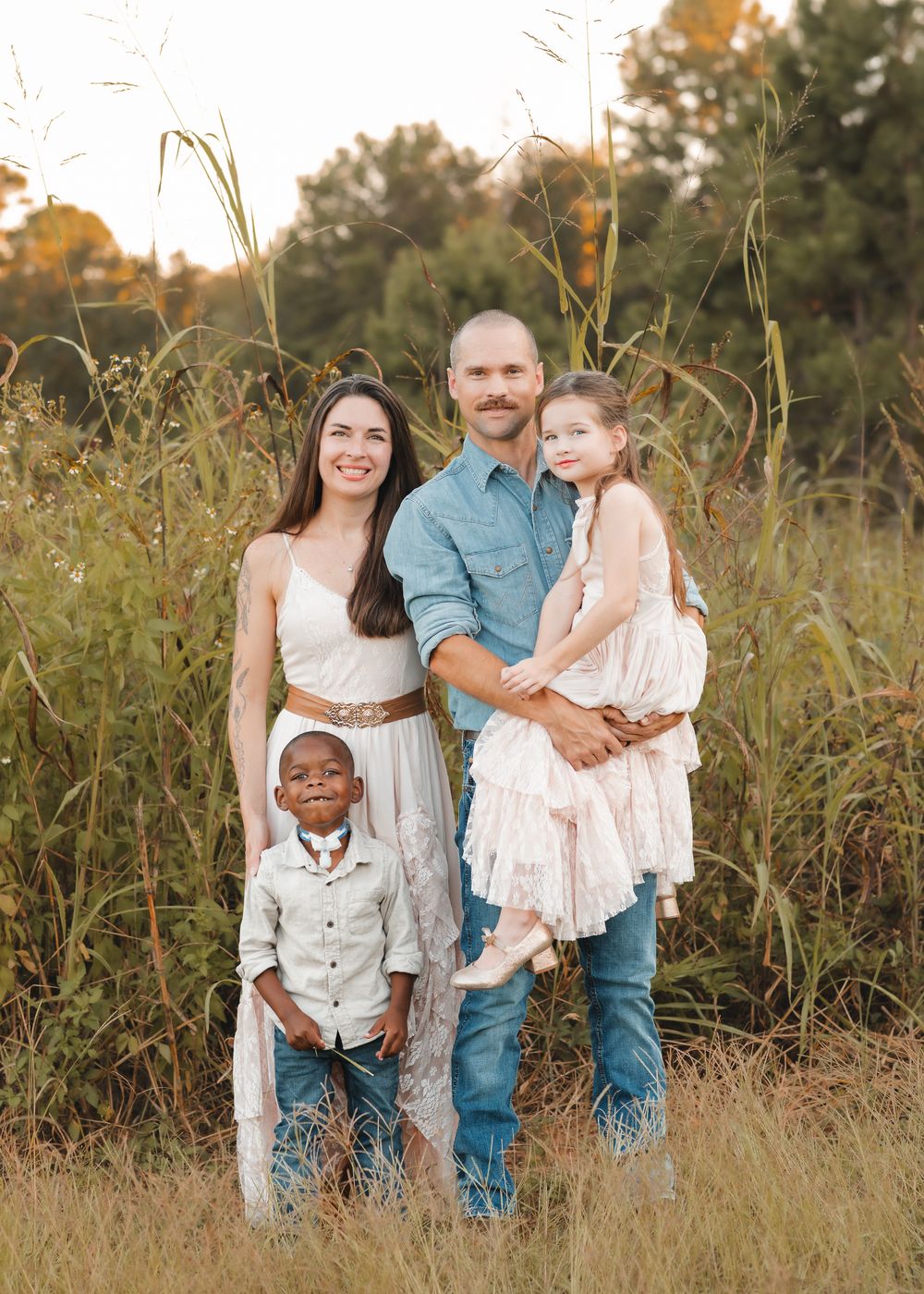 A casual outdoor family portrait session in tall grass with warm evening sunlight filtering through trees.
