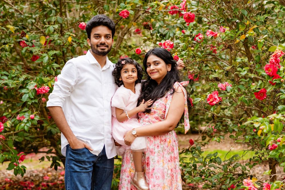 A happy South Asian family posing in a lush garden with blooming red roses, parents holding their young daughter.