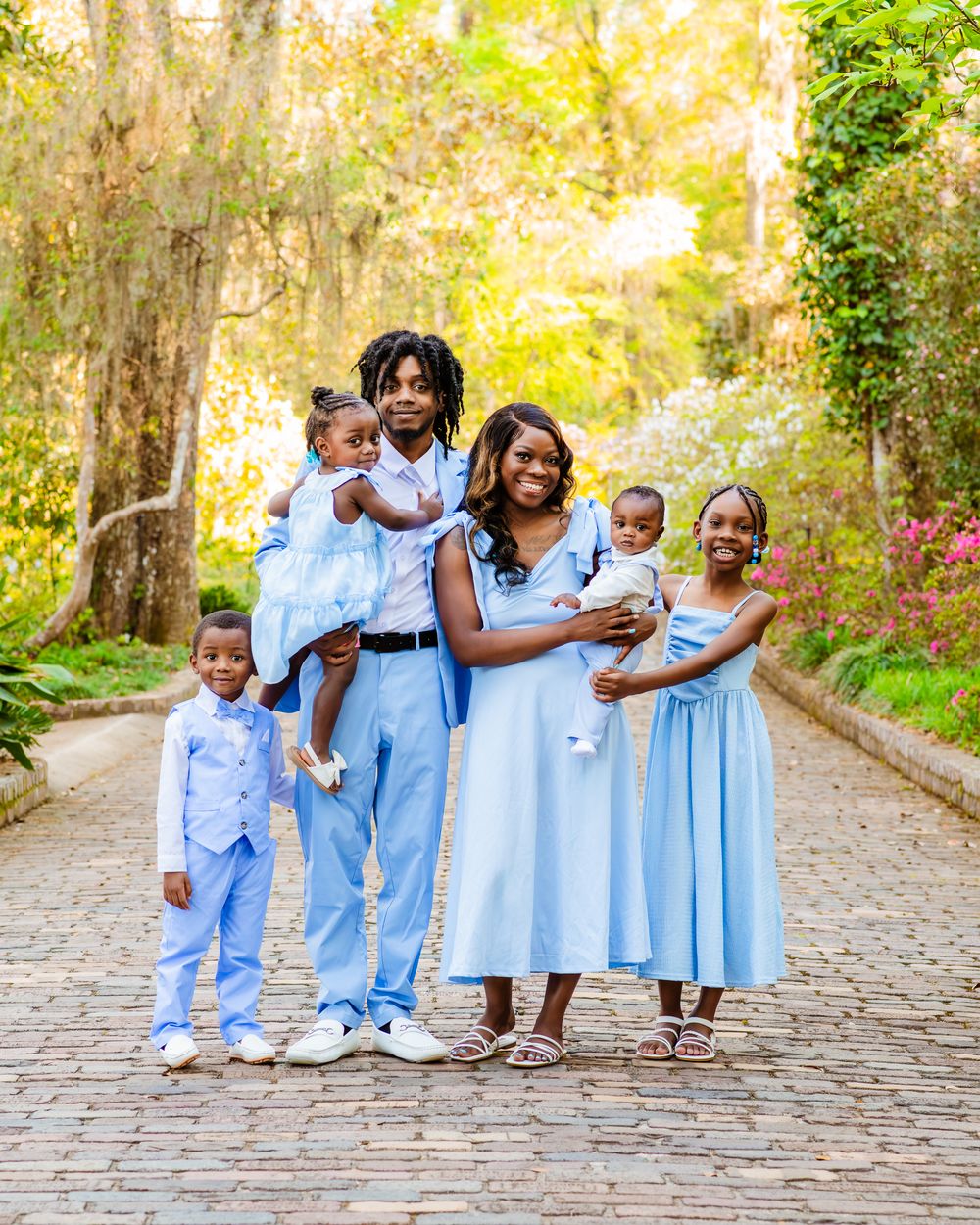 A joyful Black family of six poses in matching light blue outfits on a cobblestone path surrounded by lush greenery.