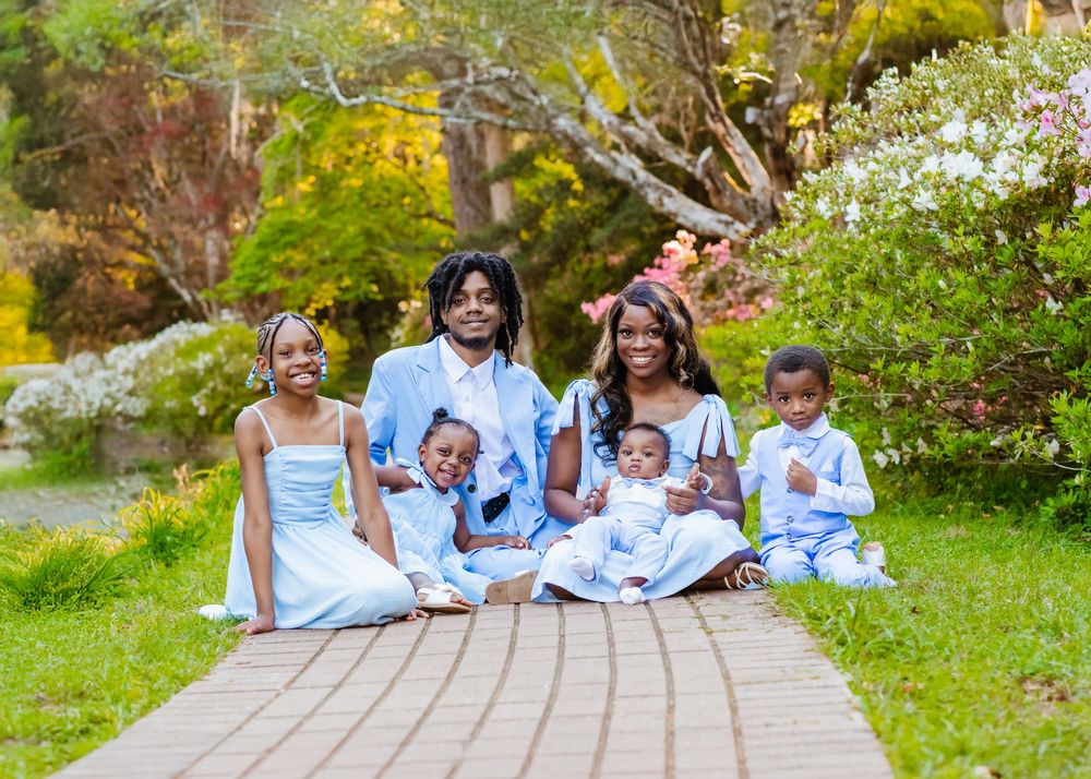 Happy family of six posing on a wooden path surrounded by lush green trees and colorful flowers.