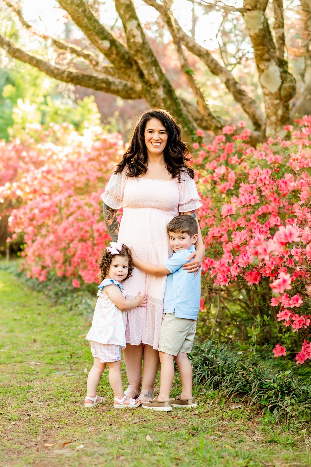 Pregnant woman in pink dress poses outdoors with two young children among blooming pink azalea bushes in spring.