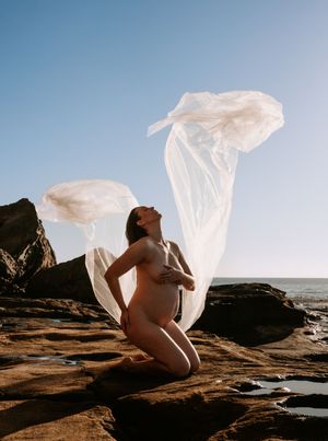 Artistic maternity photoshoot on rocky beach with flowing white fabric.