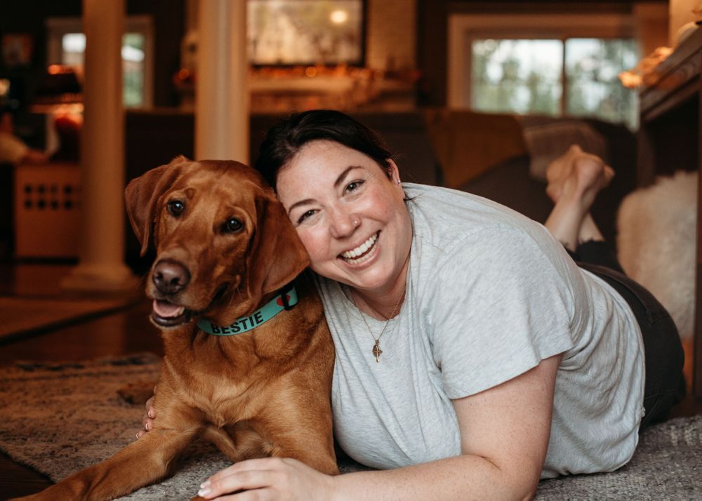 A young woman lays on the ground and snuggles up with her pooch and has a big smile while having her picture taken.