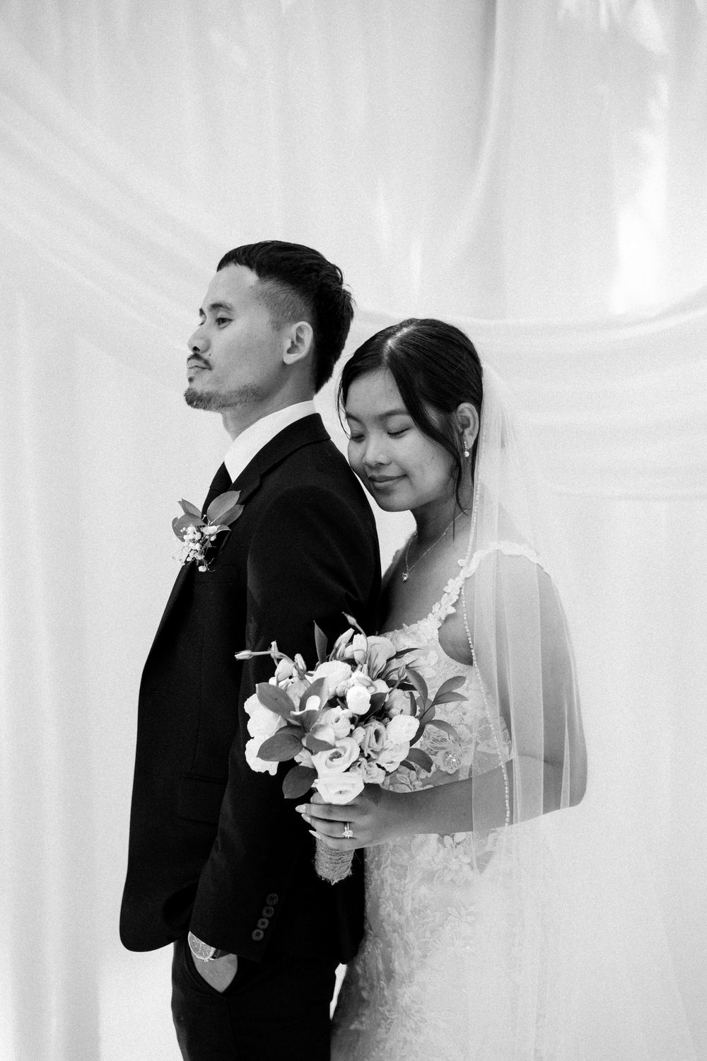Black and white wedding photo of bride holding bouquet leaning on groom in suit, elegant draping backdrop.