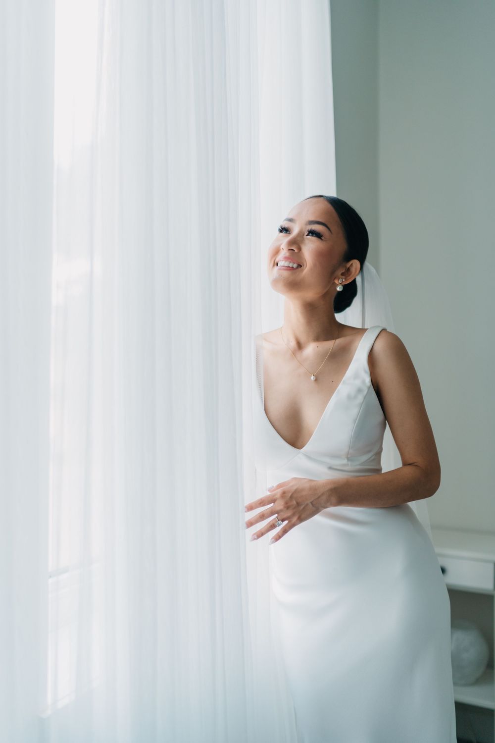 Smiling bride in white V-neck wedding gown standing by sheer curtains near a bright window.
