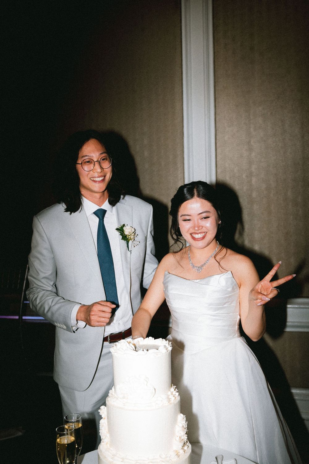 A smiling couple cuts their white tiered wedding cake, bride in strapless gown flashing peace sign, groom in gray suit.