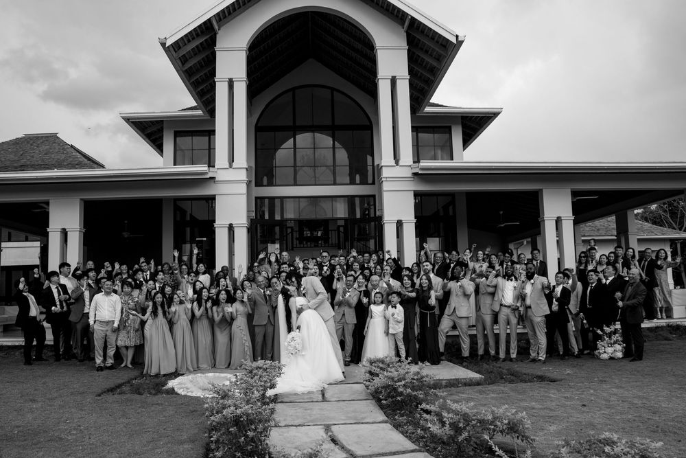 Large wedding group photo with bride and groom centered before an elegant venue, captured in black and white.
